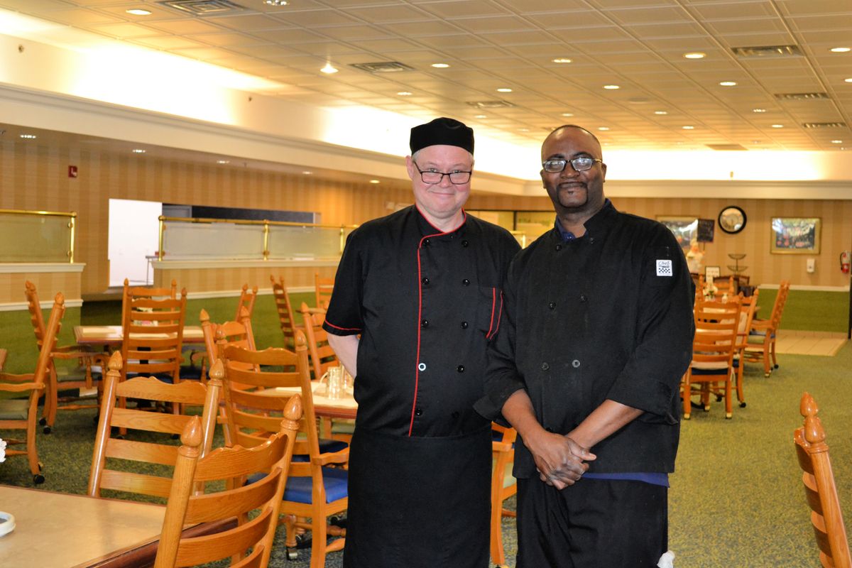 Two chefs standing in a restaurant dining area wearing black uniforms.