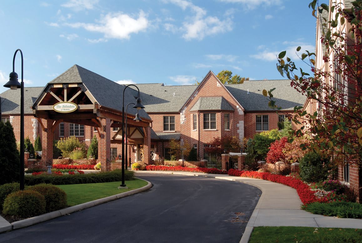 A scenic view of a brick building with a landscaped entrance and driveway under a partly cloudy sky.