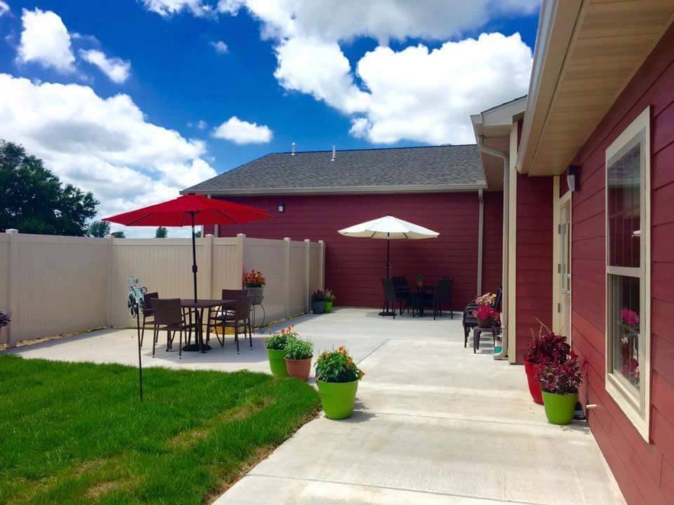 A sunny backyard patio with outdoor furniture and umbrellas, surrounded by a grassy area and a fence.