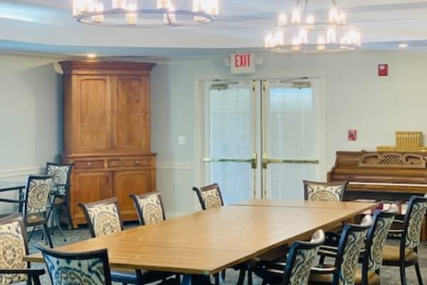 A spacious conference room with a large meeting table, surrounded by chairs and lit by elegant chandeliers. A wooden cabinet and a piano are seen in the background.