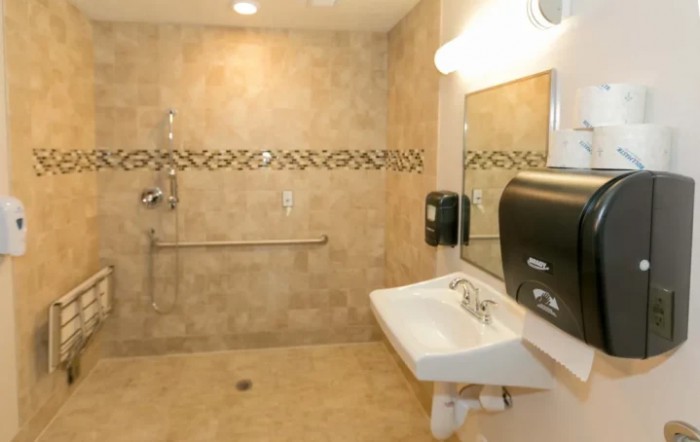 A spacious and accessible bathroom with tiled walls, featuring a sink, mirror, handrails, soap dispenser, and paper towel dispenser.