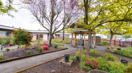 A serene garden with a gazebo, surrounded by trees and well-maintained flower beds, featuring a winding pathway.