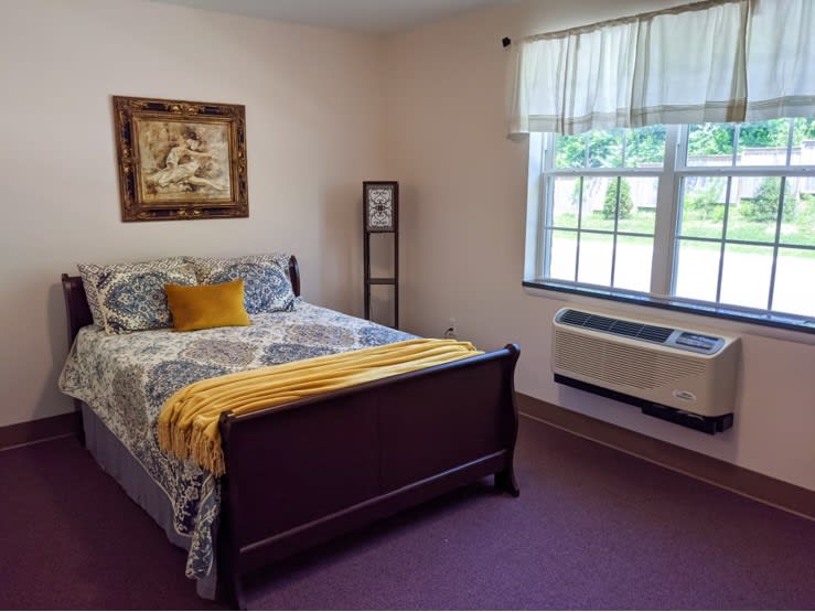 A cozy bedroom featuring a decorated bed with a patterned quilt and pillows, an air conditioner under the window, and framed artwork on the wall.