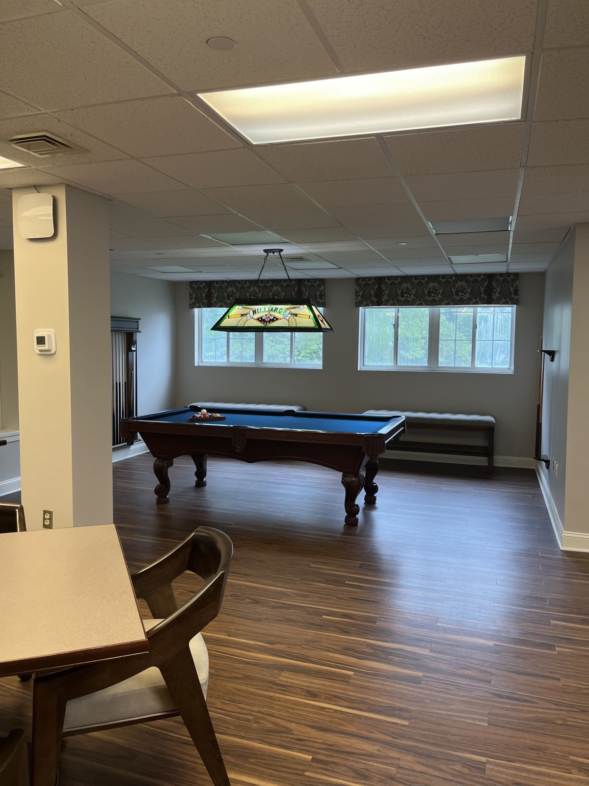 A game room featuring a pool table under a decorative light, with large windows and wooden flooring.