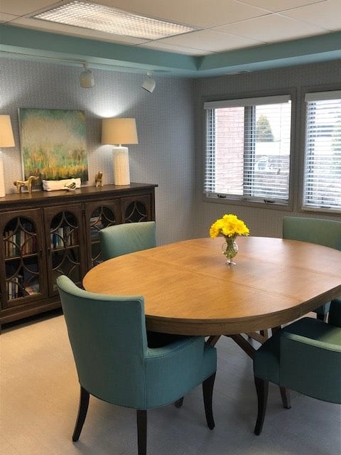 A cozy dining room with a wooden oval table surrounded by teal chairs, featuring a sideboard with lamps and landscape artwork. A vase of yellow flowers adds a touch of color.