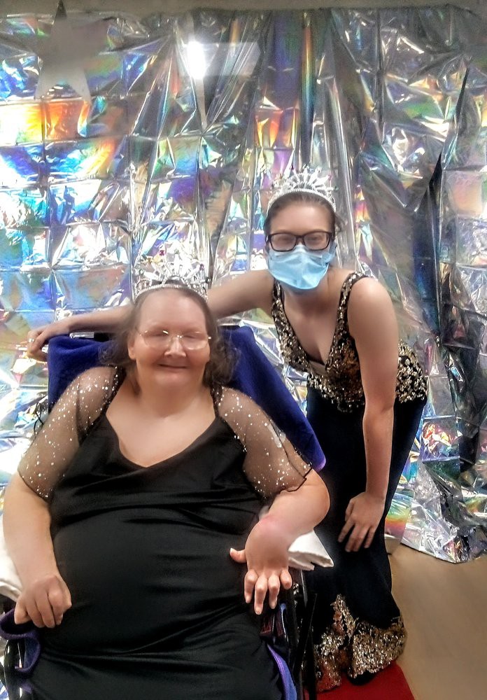 Two people at a party; one is seated in a wheelchair, and the other is standing beside them, wearing a mask and glasses.