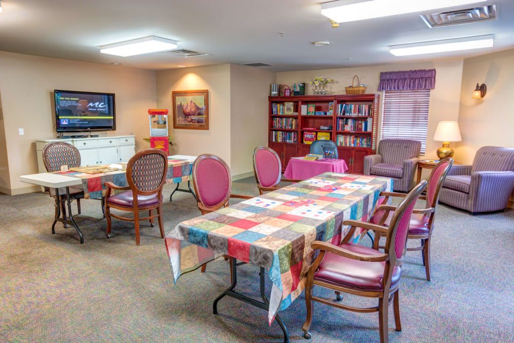A cozy community room with tables covered in colorful tablecloths, surrounded by chairs. A bookshelf and a television are visible in the background.