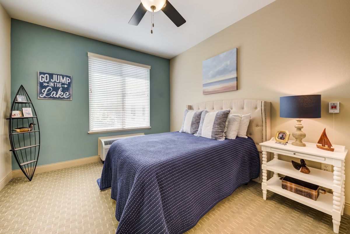 A cozy bedroom with a neatly made bed and blue bedding, featuring a nightstand, ceiling fan, and large window.
