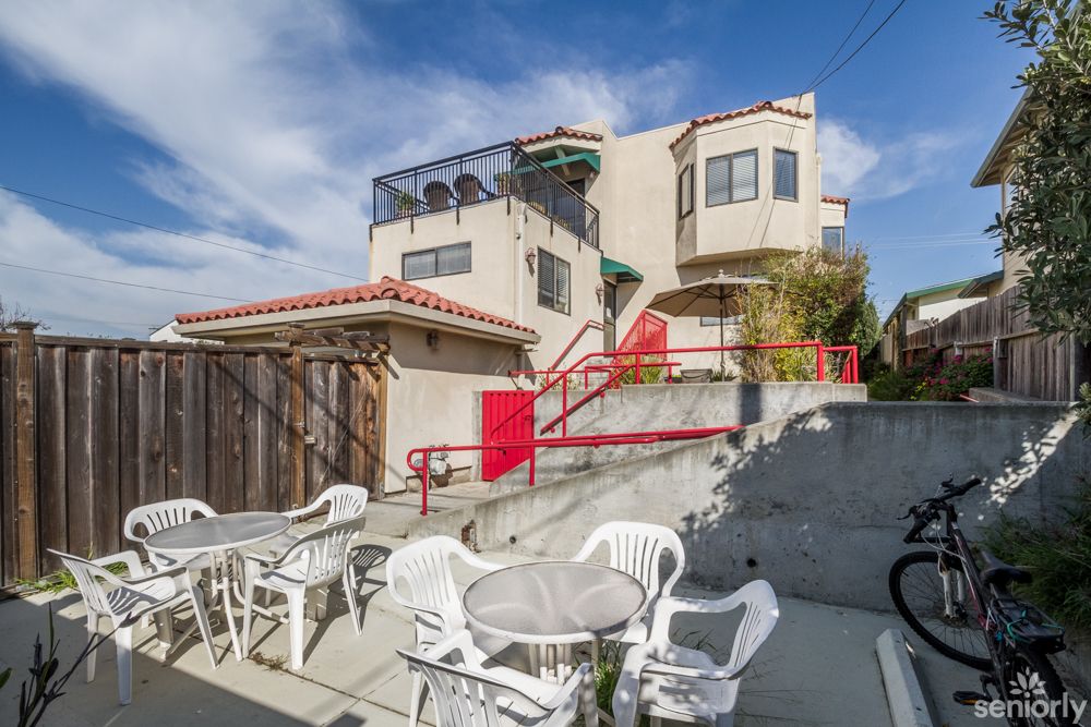 A quaint multi-story house with a patio area featuring white chairs and tables on a sunny day.