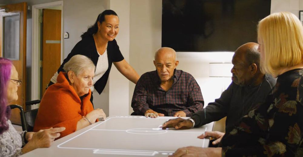 A group of elderly individuals having a discussion around a table, accompanied by a caregiver.