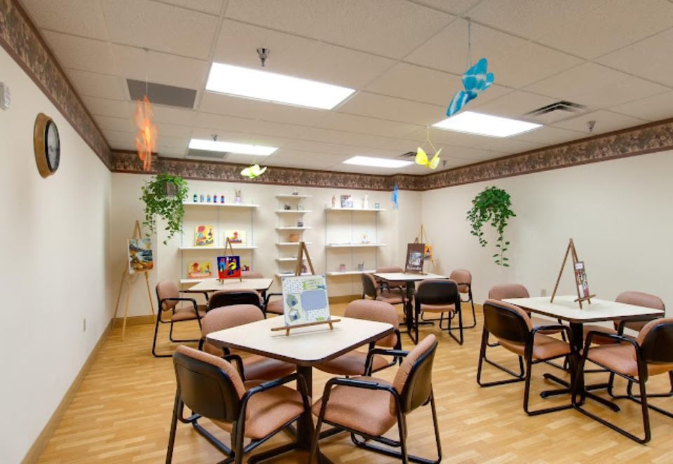A well-lit community room featuring tables and chairs, decorated with plants and art supplies on display.