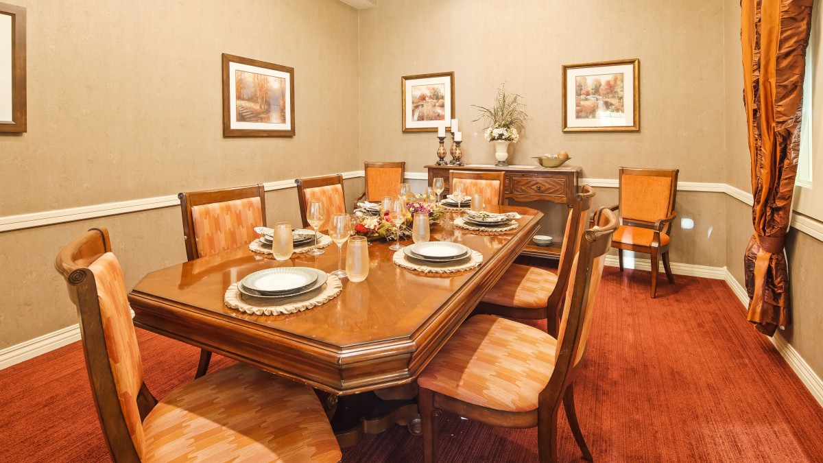 A formal dining room with a large wooden table set for dinner, surrounded by cushioned chairs and decorated with framed art on the walls.