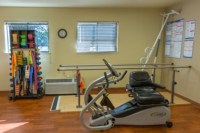 A rehabilitation therapy room equipped with a stationary bike, dumbbells, exercise balls, and parallel bars.