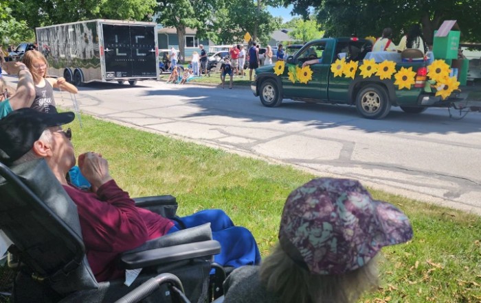 Spectators watch a parade from the sidewalk, where a decorated vehicle with sunflowers passes by.