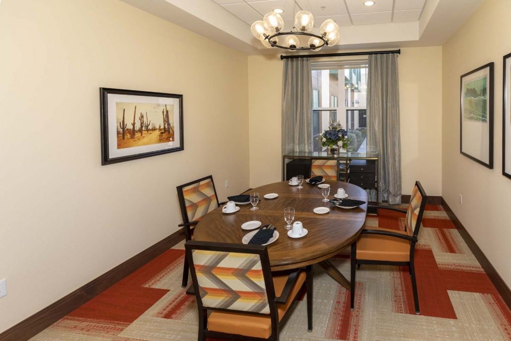 A cozy dining room featuring a wooden table set with black napkins and cups, surrounded by cushioned chairs. A chandelier hangs overhead, and a piece of artwork decorates the wall.