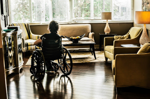 An elderly person in a wheelchair gazes thoughtfully in a cozy living room filled with natural light.