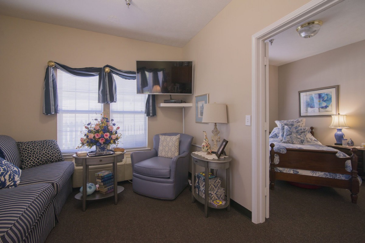 A cozy living room area with a comfortable chair, TV mounted on the wall, and decorative elements such as flowers and a lamp.