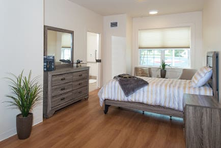 A modern bedroom with a comfortable double bed and dresser, featuring natural light from large windows.