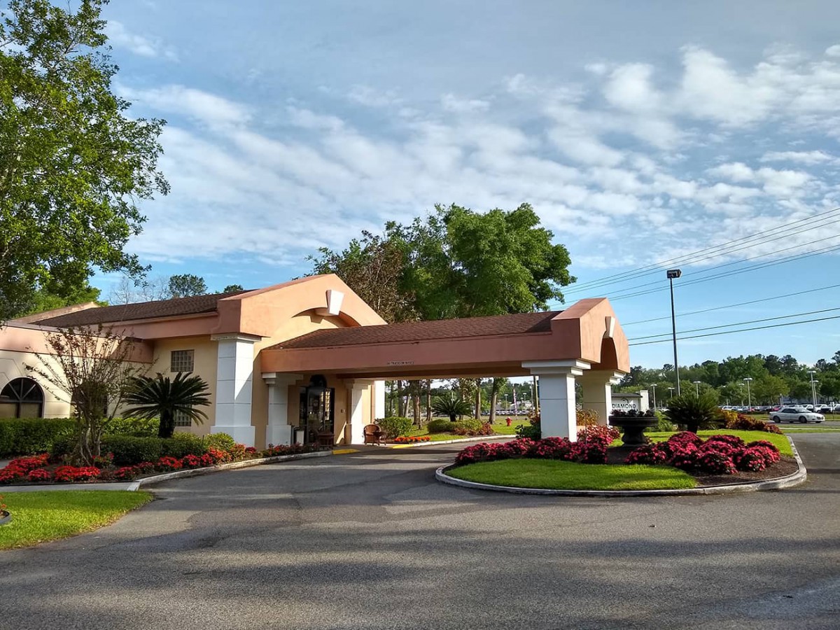 A building with a covered entrance and landscaped surroundings, featuring colorful flowers and trees under a cloudy sky.