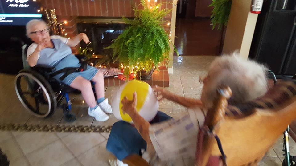 Two people playing with a beach ball indoors, one seated in a wheelchair near a fireplace.