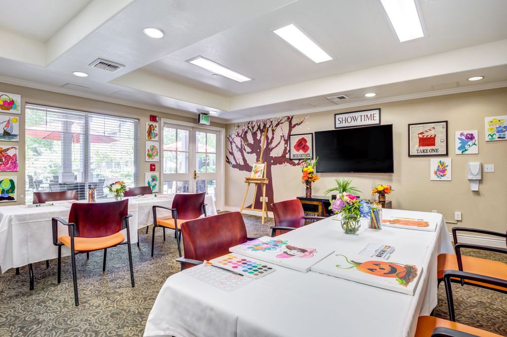 A well-lit room with tables set up for art activities, featuring colorful chairs, decorations, and a television on the wall.