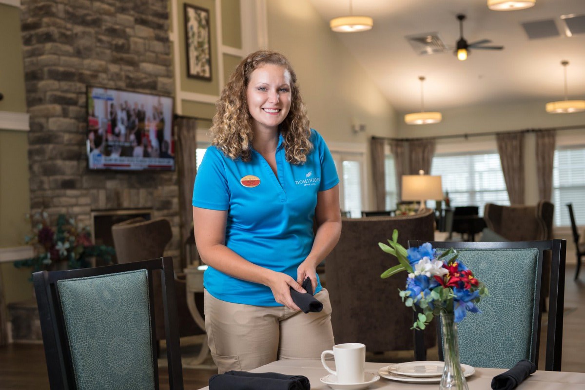 A smiling woman in a uniform setting a table in a restaurant with floral decoration.