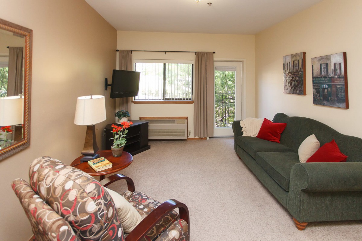 A cozy living room with a sofa, armchair, and television, featuring warm lighting and a window overlooking greenery.
