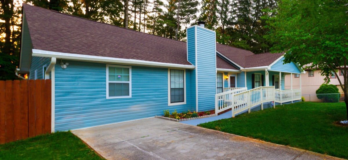 A single-story blue house with a chimney and a ramp leading to the entrance, surrounded by trees.
