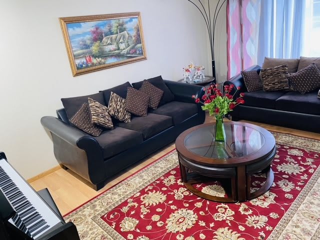 A cozy living room featuring a dark sofa set, a round coffee table with a vase of flowers, and a patterned red rug.
