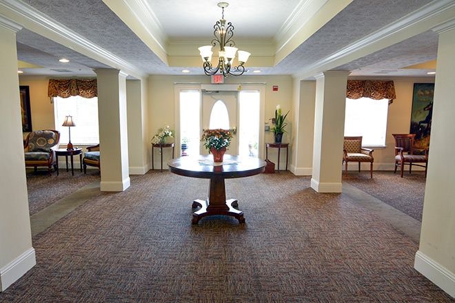 A well-lit interior space featuring a round table with floral decoration, surrounded by windows and elegant furnishings.