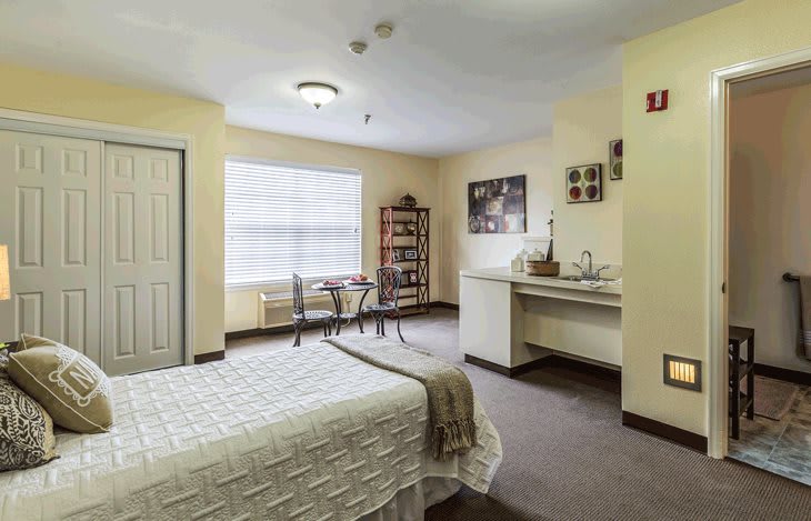 A cozy bedroom with a neatly made bed, table and chairs near the window, and artwork on the walls.