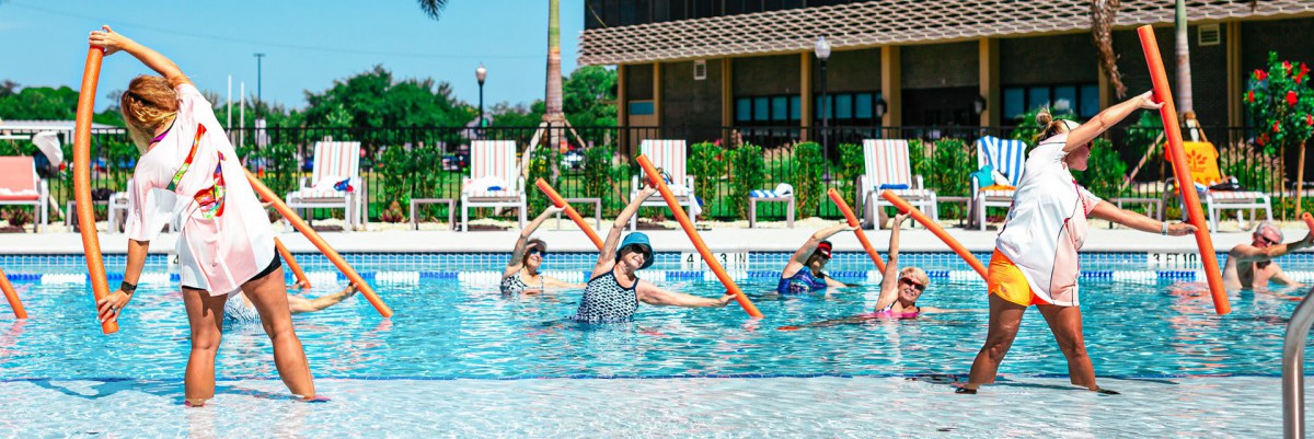 A group of people participating in a water aerobics class in an outdoor pool, using foam noodles for exercise.