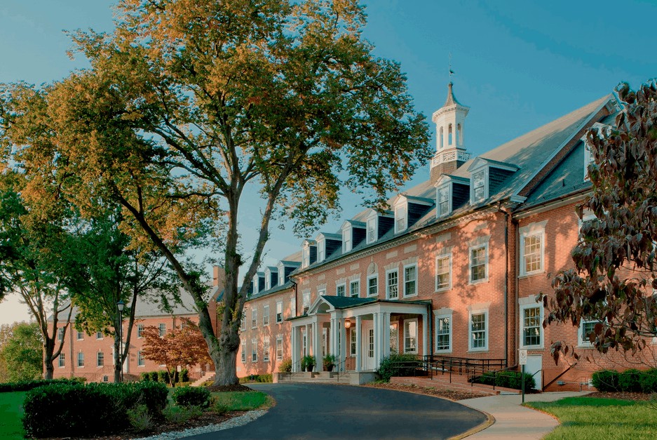 A historic brick building with a tall tree and a winding road in the foreground, set against a clear blue sky.
