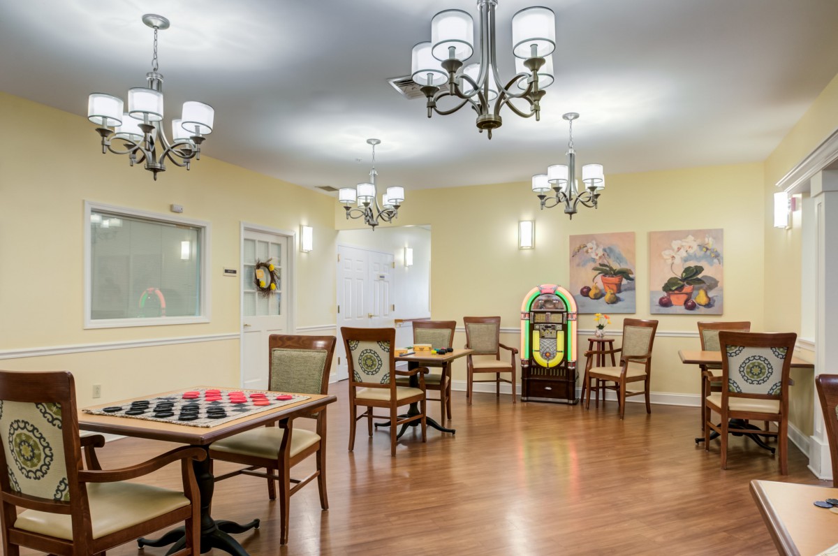 A spacious recreation room featuring tables, chairs, chandeliers, and a jukebox, with artwork on the walls.