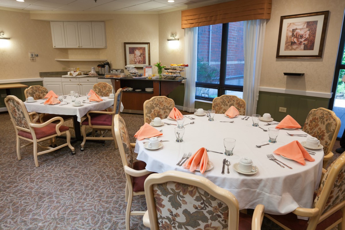 A neatly arranged dining room with tables set for a meal, featuring elegant chairs and orange napkins.