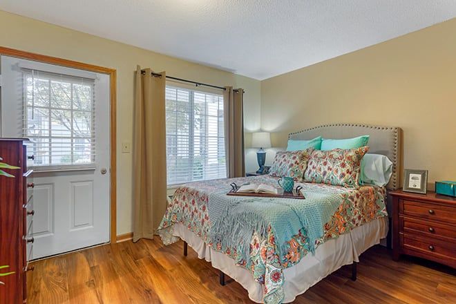 A cozy bedroom with a decorative bedspread, hardwood flooring, and natural light streaming through the window.