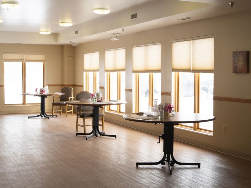 A spacious, sunlit room with tables and chairs arranged near large windows with blinds.