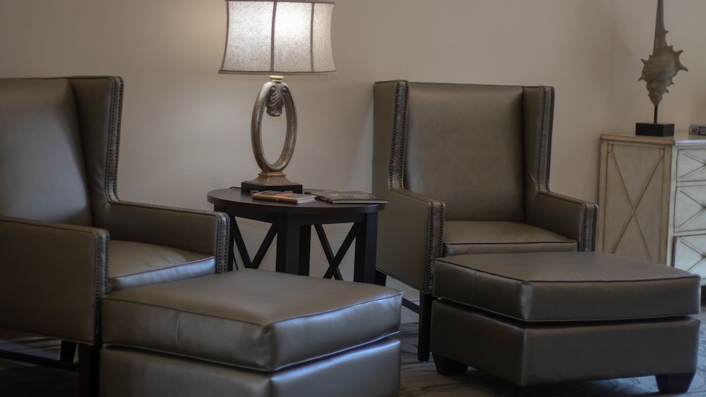 A cozy seating area with a chair, ottoman, table, and lamp.