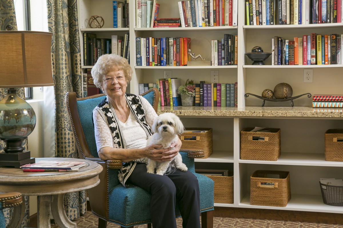An elderly woman sitting in a cozy living room, holding her small dog on her lap.