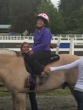 A person is enjoying a therapeutic horseback riding session with guidance from assistants.