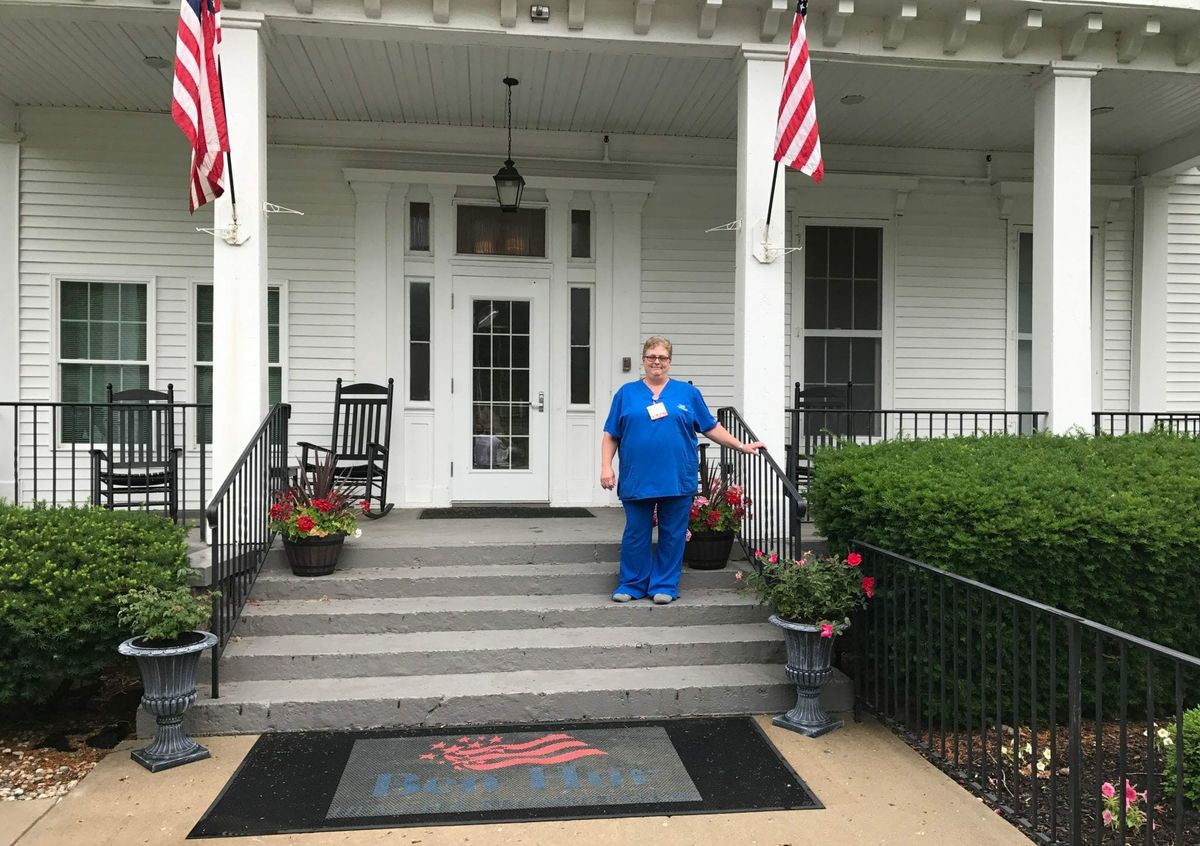 A person in a nurse's uniform stands on the steps of a building with American flags.