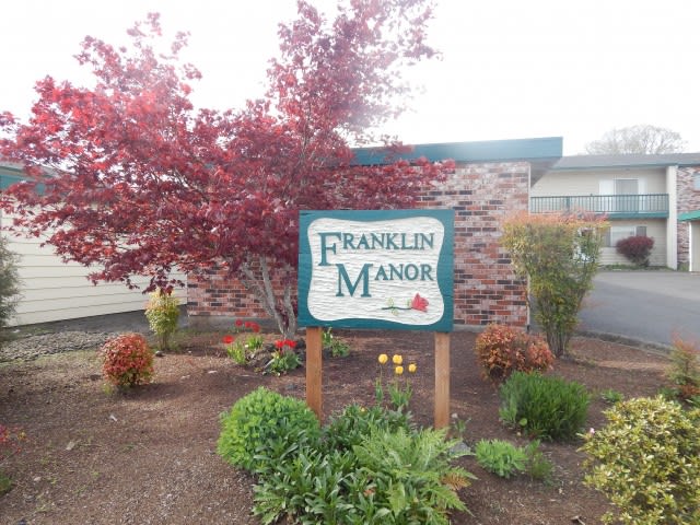 A sign reading 'Franklin Manor' stands amidst autumnal landscaping with a red-leaved tree and surrounding bushes in front of a building.