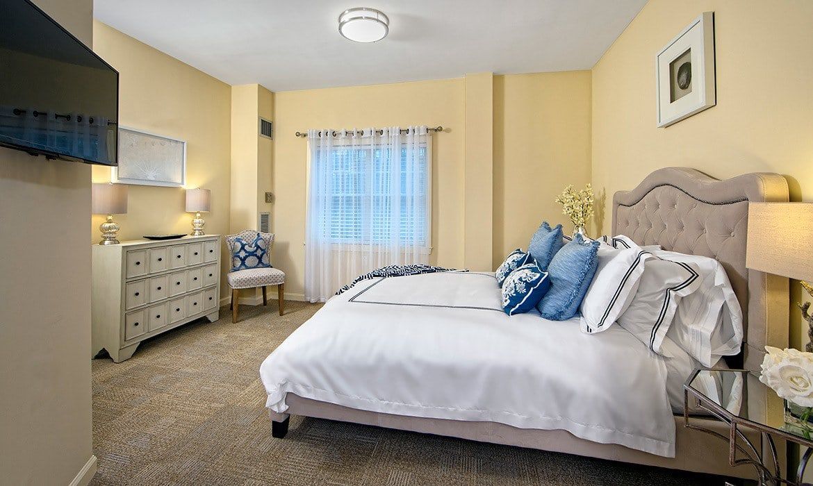 A cozy bedroom with soft yellow walls, featuring a neatly made bed adorned with blue pillows, a dresser, and a window with sheer curtains.