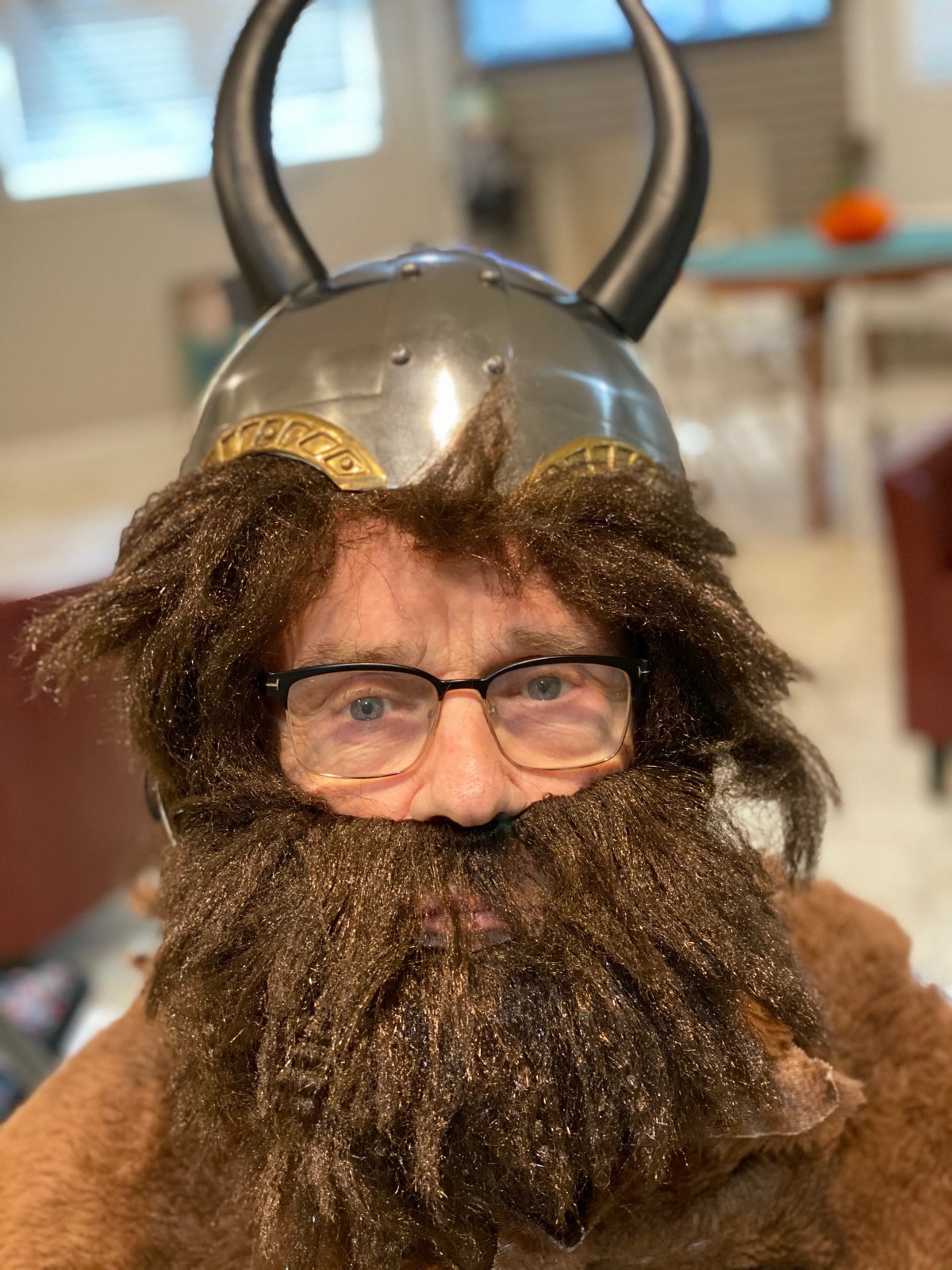 A person wearing a Viking costume complete with a helmet, beard, and glasses indoors.