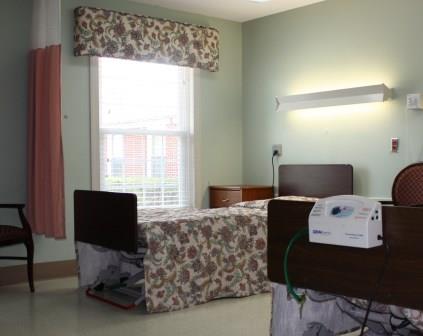 A view of a hospital patient room featuring a bed, window with floral curtains, and medical equipment.