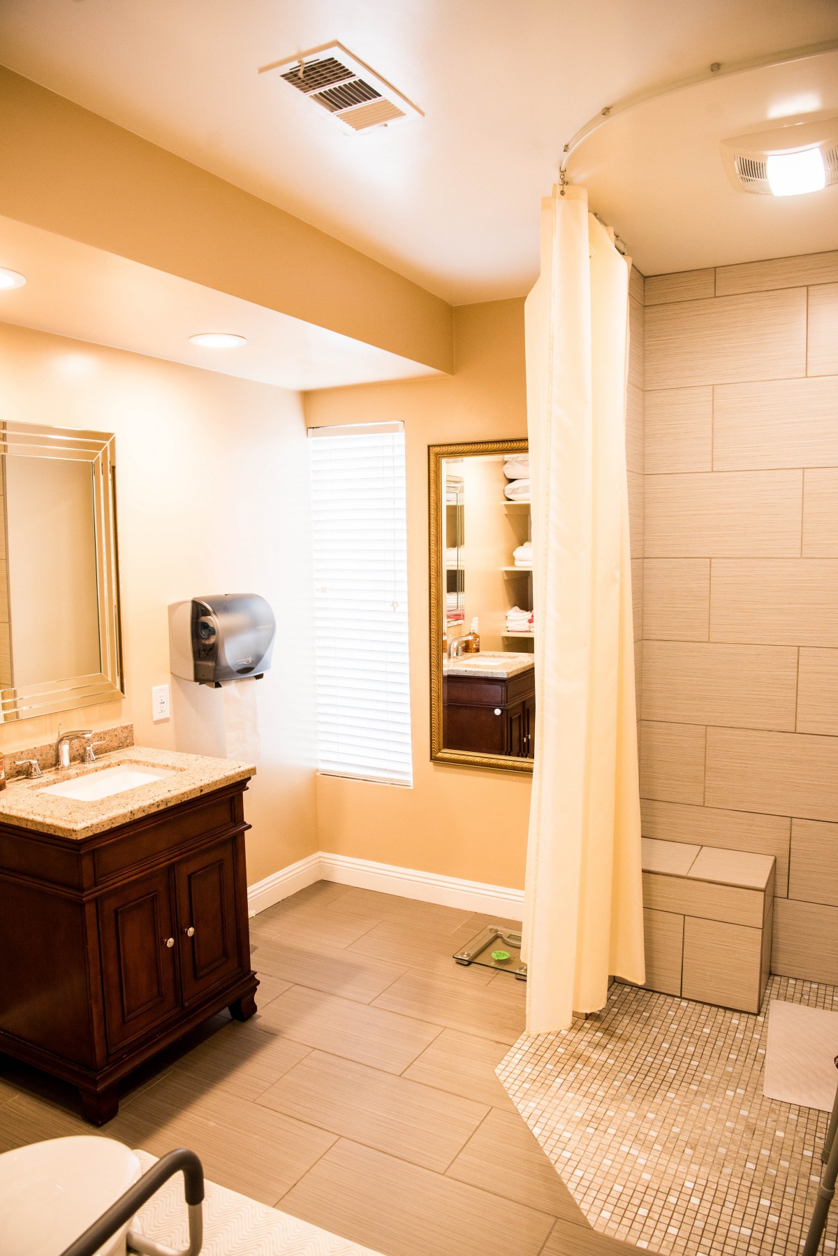 A modern bathroom with a wooden vanity, mirror, shower area, and paper towel dispenser.