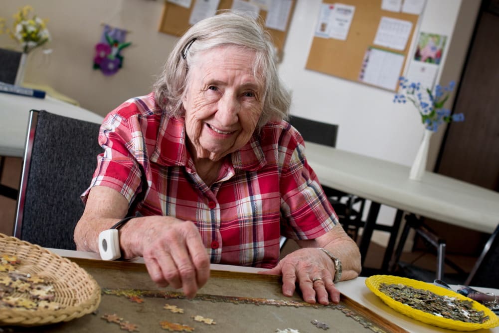 An elderly woman smiles while working on a puzzle at a table indoors.
