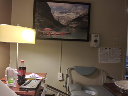 A hospital room featuring a lamp, a small table with personal items, and a landscape painting on the wall.