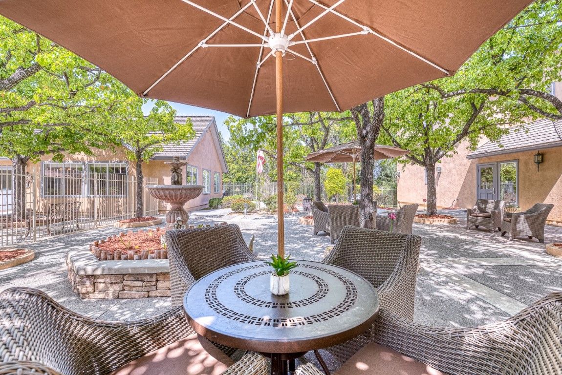 A serene outdoor patio with a table and chairs under a large umbrella, surrounded by trees and greenery.