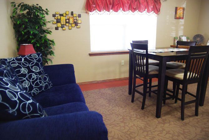 A cozy living room featuring a blue sofa, dining table with chairs, an indoor plant, and a sunlit window with red curtains.